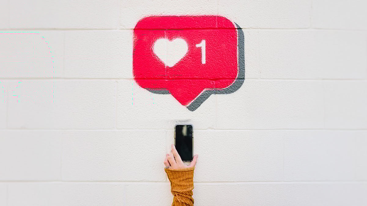 An einer weissen Wand aufgesprühtes Like-Symbol von Instagram, darunter eine Hand der Zielgruppe, die ihr Smartphone darunter hält