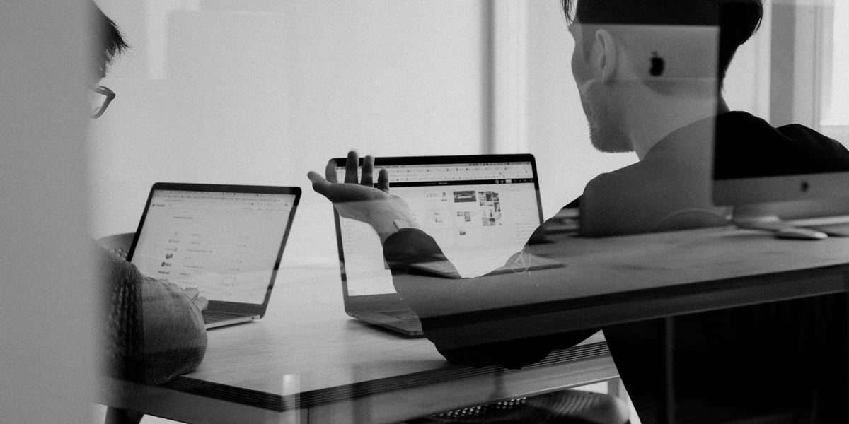 Zwei Personen arbeiten gemeinsam an Laptops in einem Büro, eine davon gestikuliert erklärend vor dem Bildschirm.